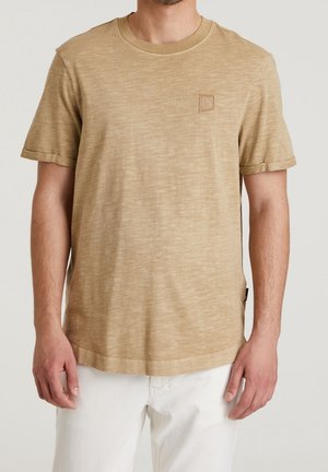 Homme portant un t-shirt beige à manches courtes avec un petit logo cousu sur la poitrine gauche, associé à un pantalon blanc.