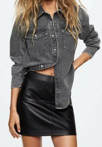 Chemise en jean grise à boutons avec deux poches poitrine, portée sur une mini-jupe en cuir noir, mettant en valeur une coupe élégante et une texture lisse.