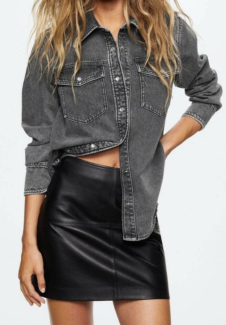 Chemise en jean grise à boutons avec deux poches poitrine, portée sur une mini-jupe en cuir noir, mettant en valeur une coupe élégante et une texture lisse.