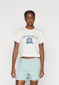 Fehér, crop fazonú póló zöld "MONACO" felirattal, lila kör alakú grafikával; világoskék rövidnadrággal kombinálva. Sima textúra, laza fazon.