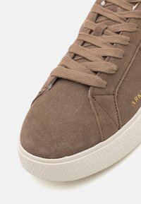 Baskets en suède marron avec lacets plats et semelle en caoutchouc blanc texturé. Caractéristiques d'un design cousu et d'un petit détail de logo doré.