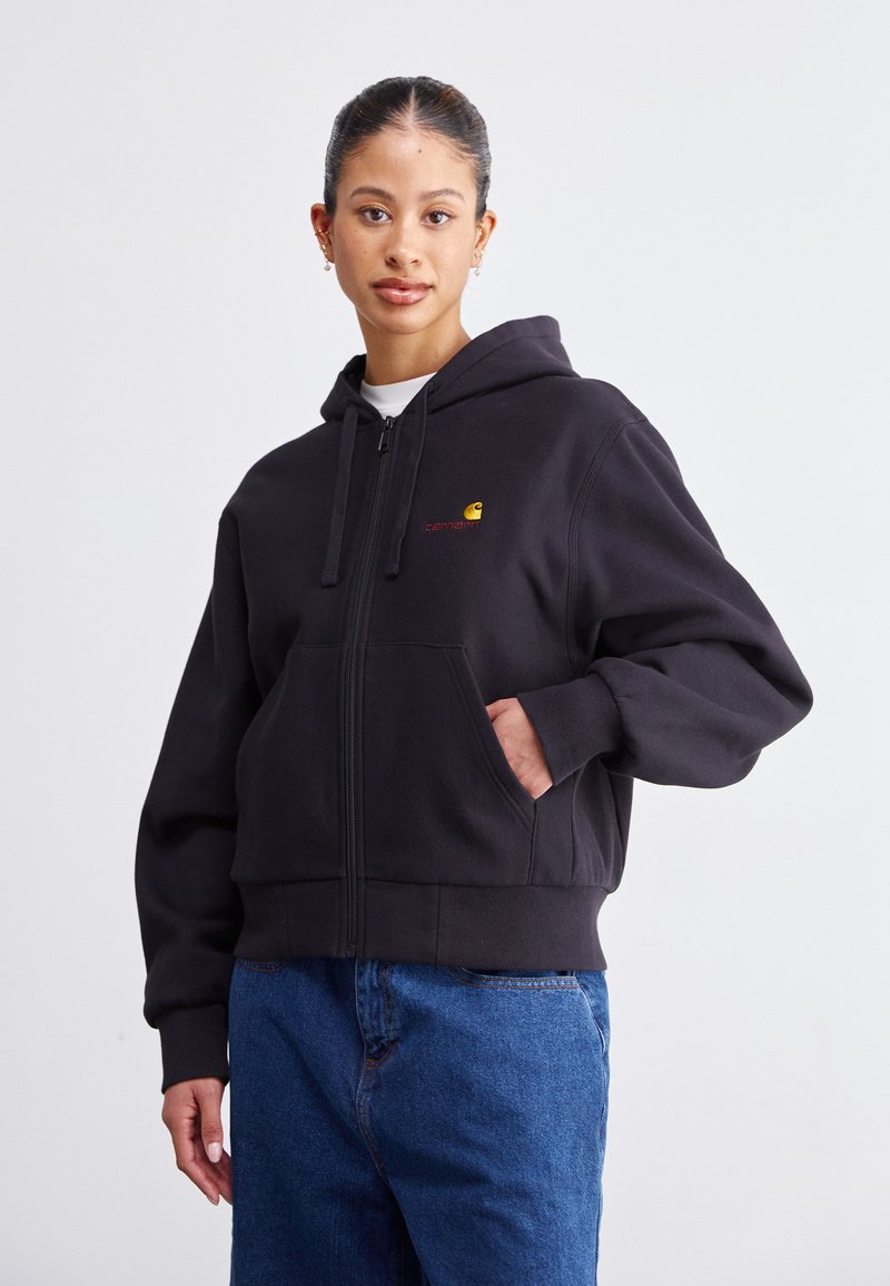 Hoodie zippé noir en tissu doux avec une poche avant, arborant un petit logo jaune et un texte rouge au niveau de la poitrine gauche.