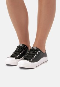 Zapatillas de lona negras con suelas de goma blancas, punta redondeada, ojetes de metal, cordones planos y logotipo en la lengüeta.