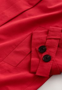 Roter Stoffjackenärmel mit einem Manschettendetail, das zwei große schwarze Knöpfe und sichtbare Nähte auf einer glatten Oberfläche zeigt.