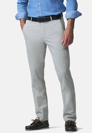 Man draagt lichtgrijze broek, een opgerolde blauwe blouse, zwarte gevlochten riem en zwarte bootschoenen met bruine veters, staand met handen gedeeltelijk in de zakken.