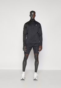 Svart långärmad atletisk zip-pullover, kombinerad med svarta shorts. Har en strukturerad tygkvalitet och vita logodetaljer. Avslutad med vita sneakers.
