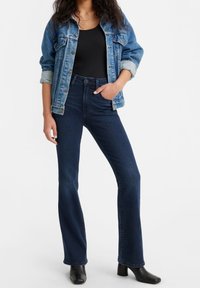 Giacca di jeans blu con un finish invecchiato, indossata sopra un top nero senza maniche. Jeans scuri a vita alta con gambe svasate e stivaletti neri.