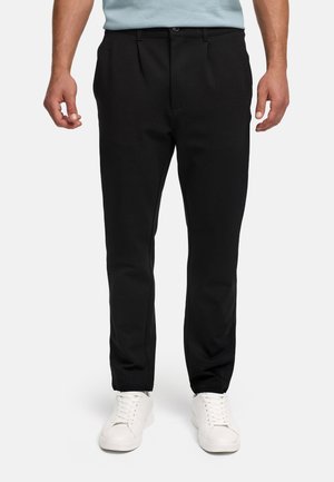 Férfi, aki fekete, testhezálló nadrágot és fehér tornacipőt visel, egy egyszínű világos háttér előtt áll.