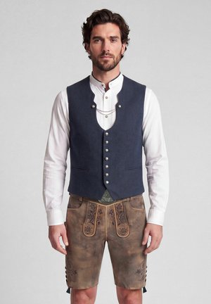 Man draagt traditionele Beierse outfit met wit hemd, donkerblauw vest met knopen en bruine leren geborduurde korte broek.