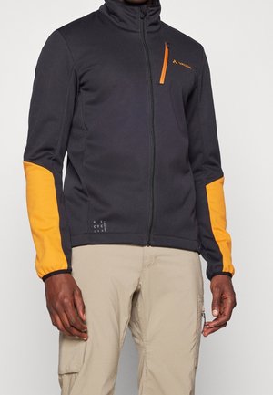 Homme portant une veste noire zippée avec des coudières orange et une fermeture éclair sur la poche poitrine, associée à un pantalon cargo beige.