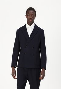 Giacca doppiopetto blu navy realizzata in tessuto testurizzato, con due tasche frontali e bottoni neri, indossata sopra una camicia bianca con colletto.