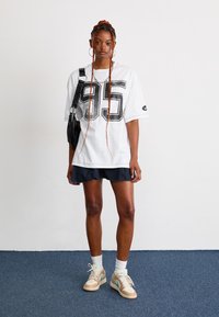 T-shirt blanc oversize avec impression noire "95", superposé à une jupe en denim bleu marine. Porté avec des baskets beiges et des chaussettes blanches. Sac à bandoulière noir.