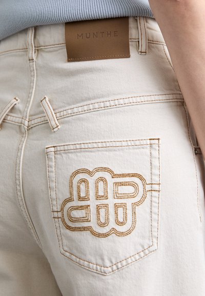 Beżowe szorty dżinsowe z wyraźnym, brązowym haftowanym logo na tylnej kieszeni. Pasek biodrowy posiada brązową skórzaną metkę.