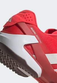 Czerwone buty sportowe z teksturowanym, dzianinowym wierzchem. Wyposażone w amortyzowaną piętę, gumową podeszwę z przyczepnością i białą paskową akcentem. Model: Adizero Ubersonic.