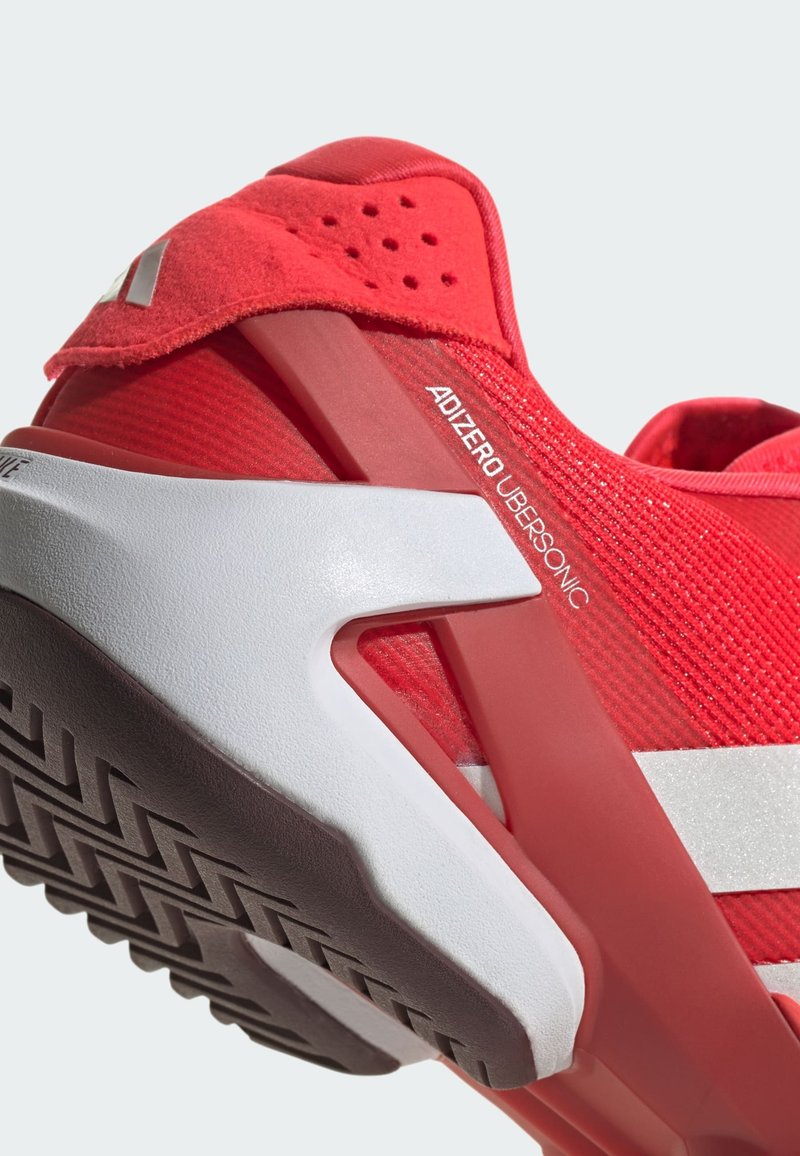 Bottine de sport rouge avec une tige en tricot texturé. Présente un talon rembourré, une semelle en caoutchouc avec traction, et une bande d'accent blanche. Modèle : Adizero Ubersonic.