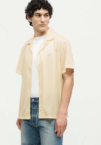 Νέος άνδρας με σγουρά μαλλιά που φορά ανοιχτό κίτρινο κοντομάνικο πουκάμισο πάνω από λευκό μπλουζάκι και μπλε τζιν, κοιτάζοντας προς τα αριστερά του.