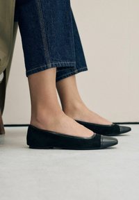Ballerine nere in suede con punta rotonda e cap-toe in pelle, caratterizzate da una texture liscia e un design minimalista. Indossate con jeans cropped.