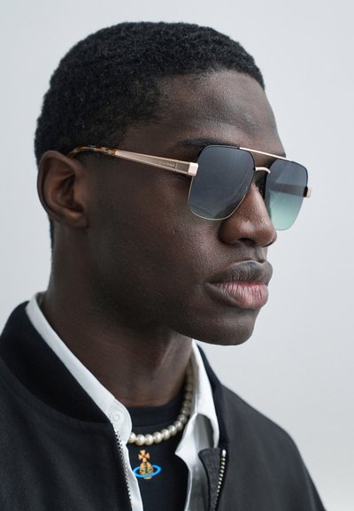 Közeli kép egy férfiról, aki négyzet alakú, színátmenetes napszemüveget, gyöngy nyakláncot, fekete dzsekit és kék-arany medállal díszített inget visel.