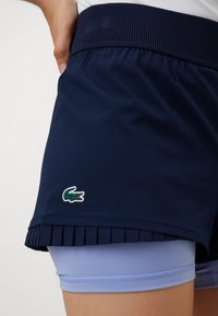 Artimiausias navy spalvos sportinių šortų su klostėmis ir šviesiai mėlynais kompresiniais šortais vaizdas, matomas ant žmogaus viršutinio šlaunies, su Lacoste krokodilo logotipu.