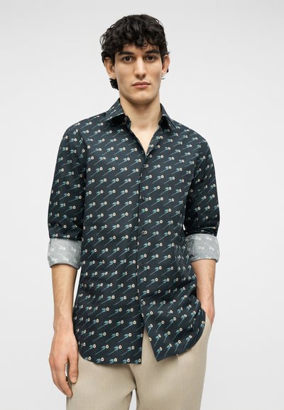 Νέος άνδρας που φοράει σκουροπράσινο πουκάμισο με μικρά λουλουδένια σχέδια, με τα μανίκια γυρισμένα και μπεζ παντελόνι, στέκεται με το ένα χέρι στην τσέπη.