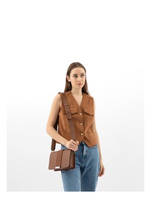 Donna con gilet marrone senza maniche e jeans blu, che porta una borsa a tracolla marrone con una cinghia larga su sfondo bianco.