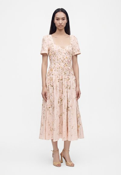 Kvinde med langt mørkt hår iført en lyserød blomsterprint midi-kjole med korte ærmer og beige høje sandaler, stående mod en hvid baggrund.