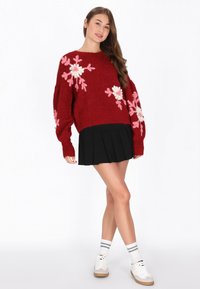 Roter Strickpullover mit floralen Mustern in Pink und Weiß, kombiniert mit einem schwarzen Faltenrock. Das Modell trägt weiße Schuhe und gestreifte Socken.