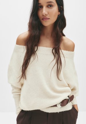 Off-shoulder cremetone strikketrøje med ribbede manchetter, stylet med højtaljede mørkebrune bukser og et bælte med sølvdetaljer.