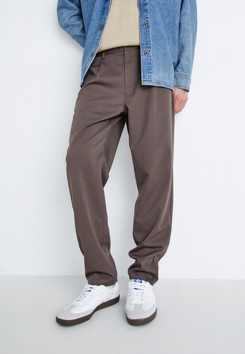 Bruine, losse broek met een gladde textuur, voorzien van een traditionele tailleband en zijzakken, gecombineerd met witte sneakers.