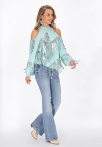 Türkiz, hideg vállú pulóver ezüst frufru részletekkel, világoskék ellensúlyozott farmernadrággal és bézs bokacsizmával párosítva.