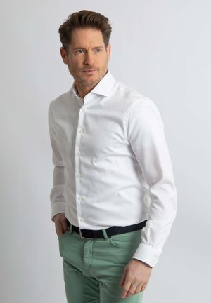 Witte lange mouwen overhemd met knopen van een licht gestructureerde stof, aangevuld met lichtgroene broek en een zwarte riem.