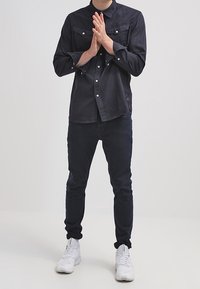 Schwarzes Hemd mit Knopfleiste und zwei Brusttaschen, kombiniert mit dunklen Jeans und weißen Turnschuhen. Das Outfit zeichnet sich durch eine schlanke Passform und ein minimalistisches Design aus.