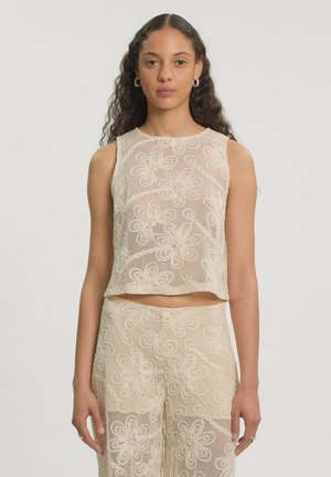 Vrouw draagt een beige kanten mouwloze top en bijpassende broek met bloemenpatronen, staand tegen een effen lichte achtergrond.