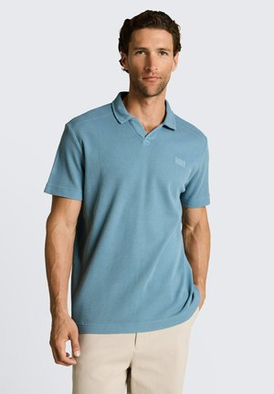 Mężczyzna z krótkimi, kręconymi włosami, ubrany w jasnoniebieską koszulkę polo z krótkim rękawem i beżowe spodnie, stoi z jedną ręką w kieszeni.