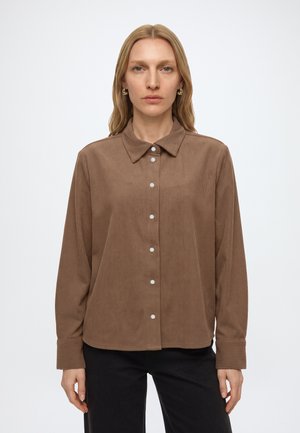 Femme aux cheveux blonds raides porte une chemise en velours côtelé marron boutonnée et un pantalon noir, debout devant un fond clair uni.