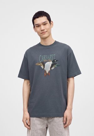 Young man wearing a dark gray T-shirt with a flying duck graphic and "Carhartt Work in Progress" text, paired with light gray pants.