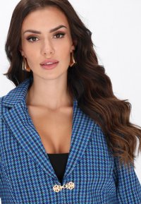 Niebieska i zielona marynarka w wzór houndstooth z klapami, z ozdobnym złotym zapięciem z przodu. Teksturowany materiał.