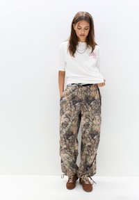 T-shirt blanche à manches courtes avec un détail graphique rose, associée à un pantalon ample à motif camouflage multicolore et des chaussures marron à lacets.