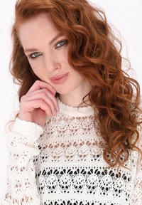 Biały sweter z koronką o skomplikowanych wzorach, z prążkowanym kołnierzykiem i mankietami oraz luźnym krojem. Modelka ma falujące rude włosy i opiera podbródek na dłoni.