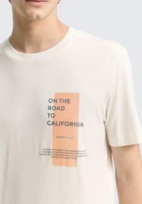 Cremefarbenes Baumwoll-T-Shirt mit kurzen Ärmeln, das einen grafischen Druck mit orangefarbener und schwarzer Schrift trägt, auf dem steht "ON THE ROAD TO CALIFORNIA."