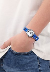 Blauwe klok met voetbalpatronen, zilveren bezel, witte wijzerplaat, zwarte cijfers en een gestructureerde band. Gied op een pols met denim en een licht shirt.