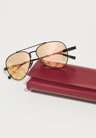 Gucci saulės akiniai su auksinėmis objektyvų tonacijomis, juodu metaliniu rėmu ir logotipo detale. Pridėtas bordo spalvos dėklas su iškilniu prekių ženklu.