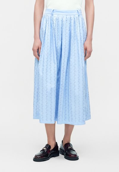 Personne portant une jupe midi bleu clair avec des motifs perforés, un haut blanc et des mocassins en cuir marron foncé.