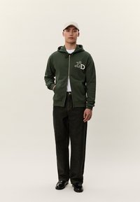 Mladý muž v zelenej mikine na zips s logom "LD", bielom tričku, čiernych nohaviciach, čiernych topánkach a béžovej čiapke, stojaci pred jednofarebným pozadím.