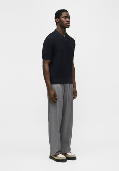 Άνδρας στέκεται φορώντας μαύρο πλεκτό πόλο με κοντά μανίκια, γκρι παντελόνι φαρδύ και μπεζ-καφέ μοκασίνια, μπροστά σε απλό φόντο.