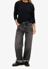 Suéter negro, jeans de pierna ancha gris con una textura desgastada. Cinturón negro con hebilla plateada. Zapatos negros con punta redonda.