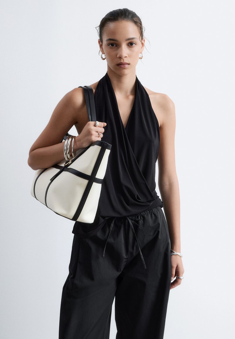 Jeune femme portant un haut noir sans manches et un pantalon, tient un sac blanc et noir sur l'épaule devant un fond uni.
