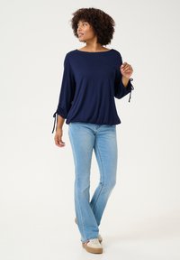 Haut bleu marine à manches longues avec des accents à nouer sur les manches, associé à un jean évasé bleu clair et des baskets beige sur fond uni.