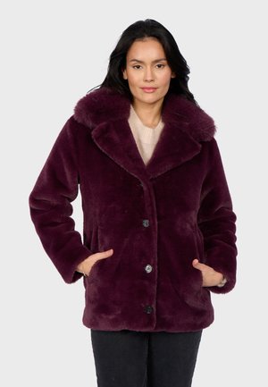 Woman wearing a plush, deep purple faux fur coat with large collar and buttons, hands in pockets, over a light beige sweater.