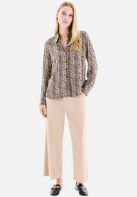 Chemise à manches longues, boutonnée, avec un motif floral en beige et noir, associée à un pantalon beige à jambes larges et des mocassins noirs avec des accents dorés.